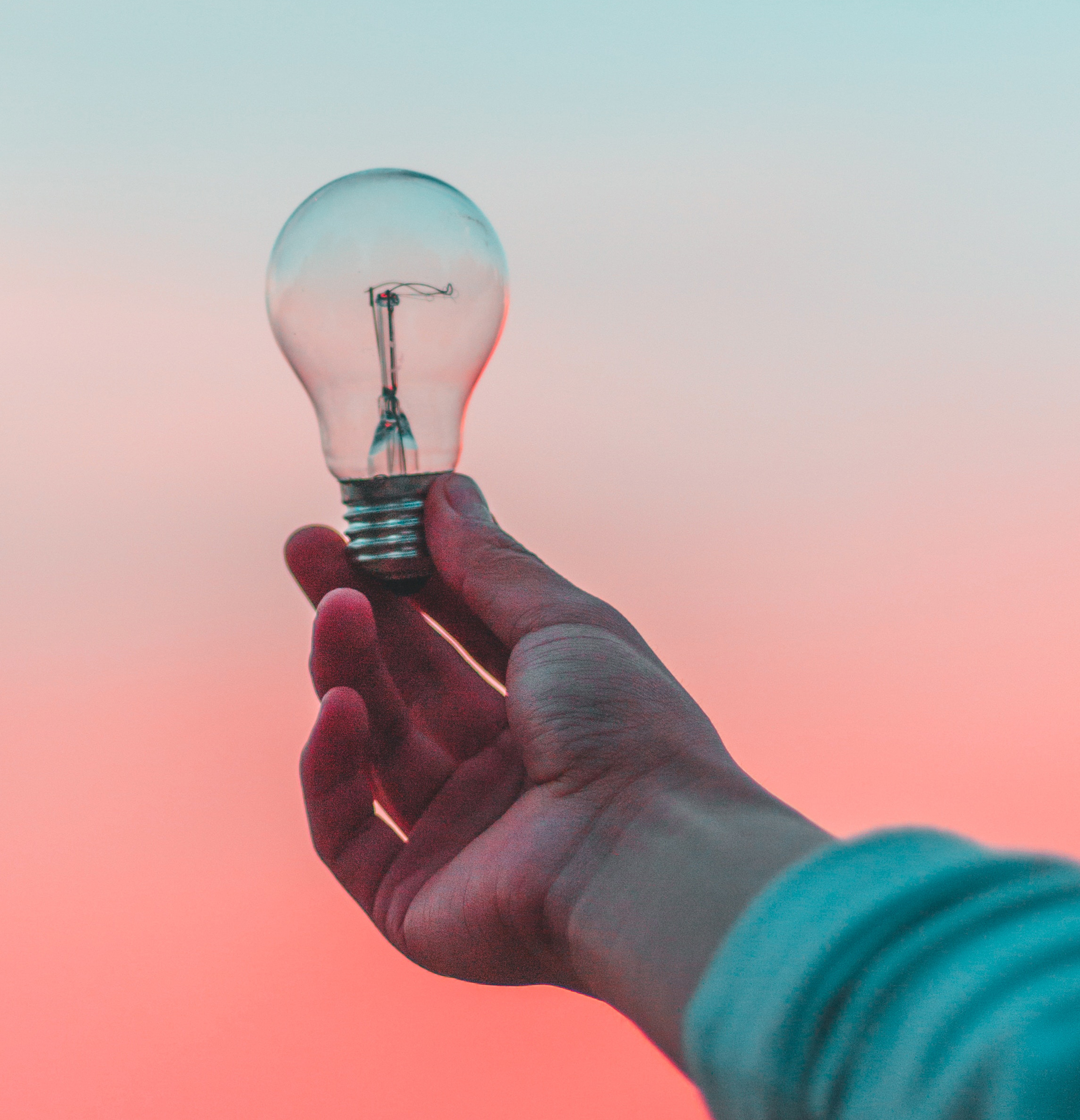 person's hand holding a lightbulb over a pink sky background