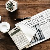 A table with the business section of the newspaper, a calculator, pad of paper, pen, cup of coffee and small plant