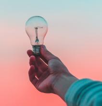person's hand holding a lightbulb over a pink sky background
